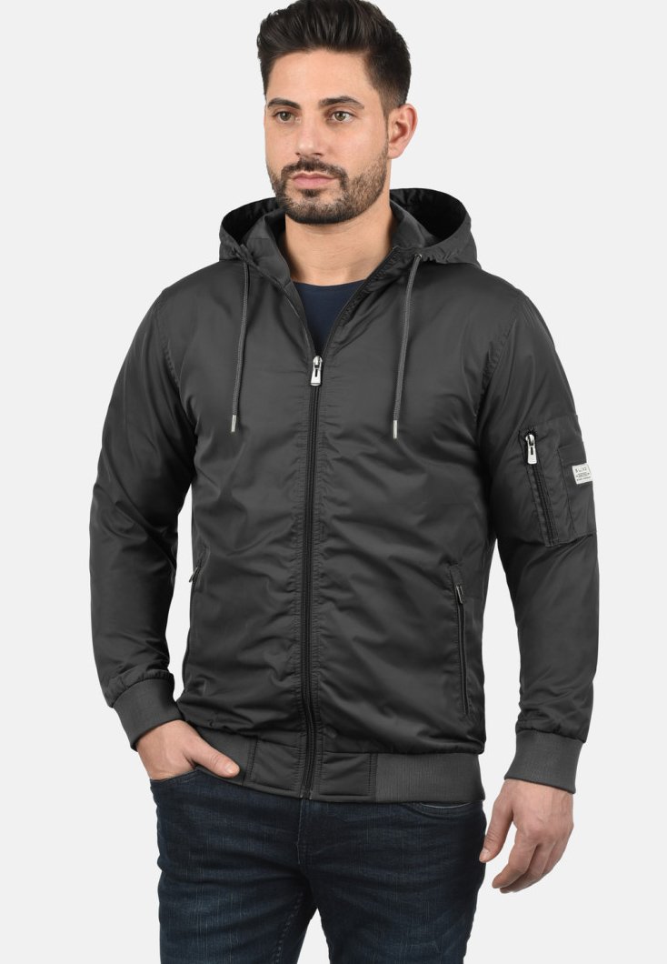 Veste à zip noire avec capuche, dotée de poignets élastiques, de poches latérales et d'une poche zippée sur le bras gauche. Tissu lisse et léger.