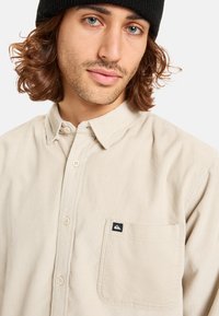 Camisa de pana beige con botón en la parte delantera, cuello puntiagudo, bolsillo en el pecho y pequeña etiqueta de logo. Textura suave con patrón de franjas verticales.