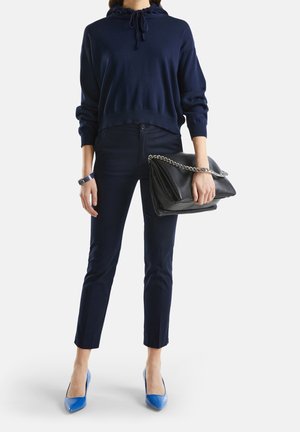 Femme portant un sweat à capuche et un pantalon bleu marine, tenant une pochette en cuir noir avec une chaîne argentée, et portant des talons aiguilles bleu vif.