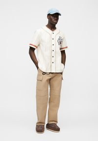 Λευκό πουκάμισο με ρίγες και κόκκινες λεπτομέρειες, με λογότυπο κλόουν, συνδυασμένο με μπεζ παντελόνι καρό και καφέ παπούτσια slip-on. Ανοιχτό μπλε καπέλο.