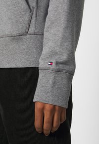 Felpa di cotone grigia con polsini a coste, con una piccola toppa con logo in rosso, bianco e blu sulla manica. Mano appoggiata su un tessuto nero strutturato.