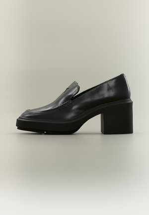 Mocassin en cuir noir avec semelle plateforme épaisse et talon bloc, avec des coutures visibles et un petit accent métallique sur la tige.