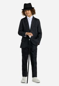 Completo black a righe nei toni del pinstripe con pantaloni abbinati, camicia bianca e cravatta, mentre tiene un bastone nero ed indossa un cappello fedora nero. Completato con sneakers bianche.