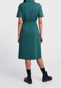 Abito camicia verde con maniche corte, vita cincinata e lunghezza al ginocchio. Indossato con stivaletti neri e calzini a contrasto. Tessuto liscio.