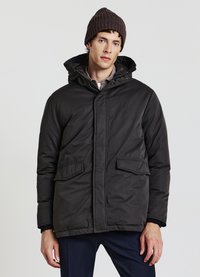 Veste matelassée sombre avec capuche, dotée d'une fermeture éclair frontale, deux grandes poches avant et une finition lisse mate. Portée avec un bonnet en tricot.