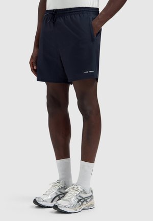 Persoon draagt marineblauwe sportshorts met de tekst "PURE PATH", witte sportsokken en zilveren hardloopschoenen, staand met handen in de zakken.