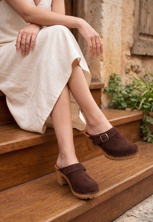 Donna in abito beige seduta su gradini di legno, che indossa zoccoli in camoscio marrone scuro con tacco in legno spesso e fibbie laterali.