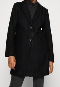 Manteau en mélange de laine noire avec un design ajusté, des revers crantés, deux boutons, des poches avant et une finition légèrement texturée.