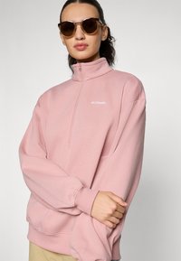 Felpa rosa in poliestere con colletto alto e zip a quarto, caratterizzata da spalle scese e un piccolo logo bianco sul petto.