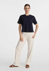 Granatowa bawełniana koszulka z krótkim rękawem zestawiona z kremowymi lnianymi spodniami. Model stoi z rękami na biodrach, w brązowych sandałach.