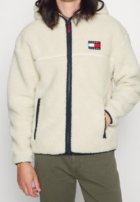 Chaqueta de forro polar color crema con cremallera frontal, ribete azul marino y un pequeño parche de la marca en el lado izquierdo del pecho con acentos rojos y verdes.