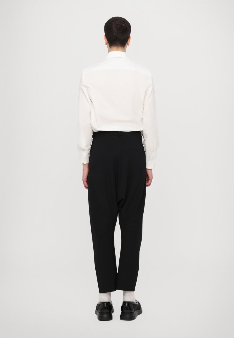 Camicia bianca a maniche lunghe e pantaloni neri attillati. Scarpe nere con suola spessa. Sfondo semplice, focalizzato sulla vestibilità e i dettagli dell'outfit.