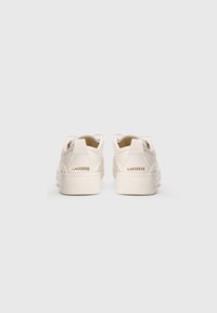 P�ár off-white tenisek Lacoste zobrazených ze zadní strany, zdůrazňující logo na patní části a čistou gumovou podrážku na jednobarevném pozadí.