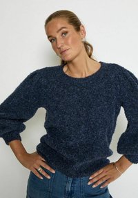 Navy gestrickter Pullover mit Ballonärmeln, Rundhalsausschnitt und geripptem Saum. Kombiniert mit hellblauer Jeans, die eine lockere Passform zeigt.