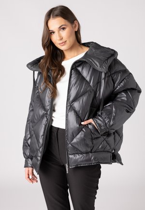 Zwarte gewatteerde jas met een capuchon, met een diamanten quiltontwerp, glanzende uitstraling en zijzakken; gedragen over een witte top en zwarte broek.