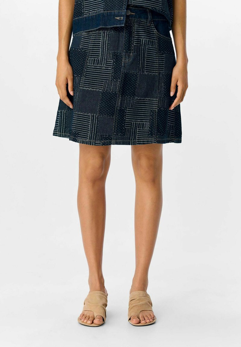 Jupe en denim avec un design en patchwork présentant des textures et motifs variés en bleu foncé, associée à des sandales à enfiler beige.