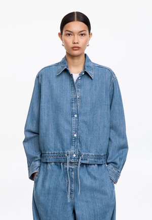 Kobieta o prostych ciemnych włosach nosi oversize'ową jasnoniebieską kurtkę dżinsową i dopasowane spodnie, stoi na tle białego tła.