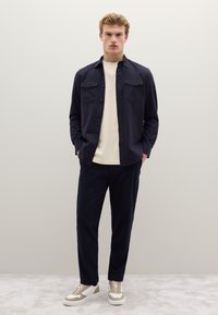 Marineblauw overhemd met knopen en twee borstzakken, daaronder een crème t-shirt, gecombineerd met donkere broek en sneakers met beige en witte accenten.