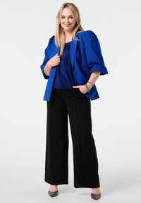 Koningsblauwe blazer met pofmouwen, gecombineerd met een glanzende blauwe top en losse zwarte broeken met zijaccenten. Zwarte schoenen met een gestructureerd oppervlak.