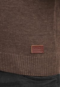 Brun strikket genser med ribbestrikket nederkant, med en strukturert stoff og et merkebadge i lær på venstre nedre hjørne.