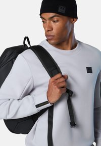 Zaino nero con spallacci regolabili, indossato sopra una felpa grigio chiaro. Etichetta con logo ben visibile sulla manica, che mostra linee di design pulite.