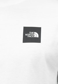 Λευκό βαμβακερό μπλουζάκι με μαύρο τετράγωνο λογότυπο με στρογγυλεμένες άκρες, που εμφανίζει το κείμενο "THE NORTH FACE" και ένα καμπυλωτό διακοσμητικό στοιχείο.