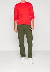 Pull en tricot rouge à manches longues, col rond, associé à un pantalon vert olive. Des baskets blanches complètent le look. Détail minimaliste du logo visible.