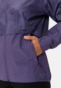 Lila wasserdichte Jacke mit Reißverschluss vorne, elastischen Bündchen, Seitentaschen und einem Logo. Glatte Textur mit leicht glänzendem Finish.