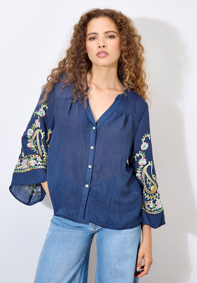 Marineblaue Bluse mit langen Ärmeln, die mit floralen Stickereien in Rosa, Grün und Gelb verziert ist. Mit Knopfleiste und aus leichtem Stoff.