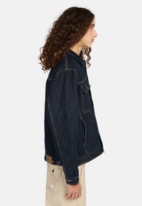 Giacca in denim blu scuro con colletto a punta, tasche frontali e cuciture a contrasto. Presenta una vestibilità rilassata e un orlo leggermente curvo.