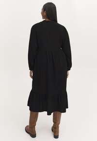 Rochie neagră cu mâneci lungi, talie strânsă, tiv volănaș și textură netedă, asortată cu botine maro până la genunchi.