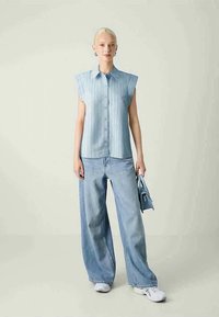 Camisa de mangas curtas, azul claro, com riscas verticais e fecho frontal de botões, combinada com jeans largos azul claro e ténis brancos.