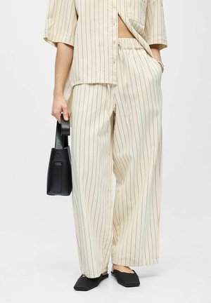 Person iført cremefarvede bukser med brede ben og med striber og en tilsvarende kortærmet skjorte, der holder en sort håndtaske og har sorte flade sko.