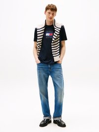 Gilet a righe beige in maglia sopra una t-shirt blu navy con logo, abbinato a jeans in denim azzurro chiaro e mocassini neri. Outfit semplice e casual.