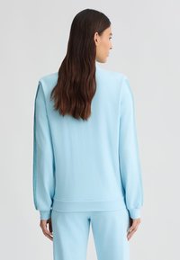 Felpa azzurra chiara con maniche lunghe, polsini a costine e dettagli in rete laterali. Materiale soffice e vestibilità comoda, abbinata a pantaloni coordinati.