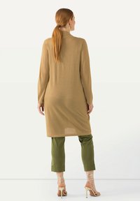 Tunique en tricot à manches longues en or métallique, associée à un pantalon slim olive. La mannequin porte des talons nus à lanières. Design simple, fente sur le côté.