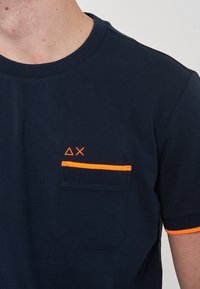 T-shirt navy con collo rotondo a coste, rifinitura arancione sulla tasca e dettagli grafici sulla tasca del petto. Maniche corte con polsini arancioni.