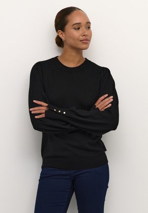 Maglione nero a maglia con maniche lunghe e voluminose e dettagli in bottone dorato sui polsini. Abbinato a jeans fitted blu scuro. Design semplice e moderno.
