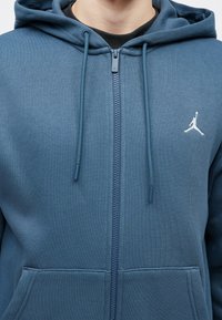 Sweat à capuche zippé bleu en tissu doux avec une poche kangourou, une capuche à cordon de serrage ajustable et un logo blanc sur la poitrine.