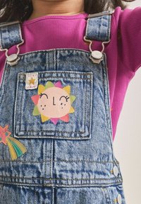Pantalones de mezclilla tipo peto con un bolsillo frontal que presenta un colorido diseño de sol y un pequeño parche de estrella; se llevan sobre una camiseta rosa de canalé.