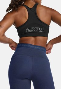 Črn športni modrček z rdečimi hrbtom, okrašen z ribasto teksturo in belim logotipom "2XU". V kombinaciji z visokim modrim leggingom.