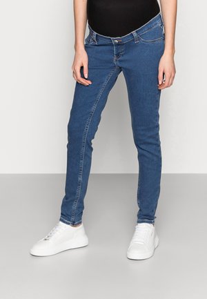 Personne portant un jean bleu slim et des baskets blanches, debout devant un fond uni.