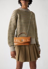 Sac à main en cuir tan avec un design incurvé et un accent en forme d'abeille dorée, doté d'une bandoulière chaîne. Porté avec un pull rayé et une jupe plissée marron.