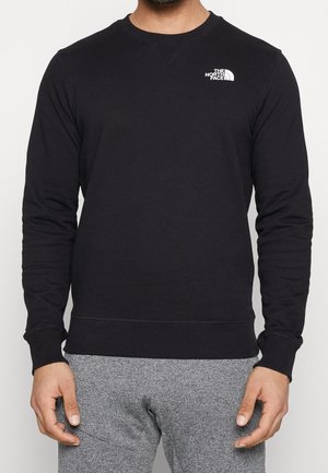 Mężczyzna w czarnej, długiej bluzie z okrągłym dekoltem z białym logo "The North Face" na piersi oraz szarych, teksturowanych spodniach.