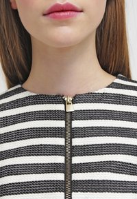 Czarno-biały materiał w paski o teksturowanym wykończeniu, z widocznym złotym zamkiem błyskawicznym przy dekolcie. Brak dodatkowych wzorów czy zdobień.