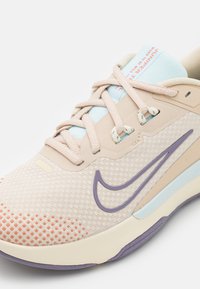 Chaussure de sport beige clair avec une surface texturée, accents violets et une languette bleu clair. Présente un dessus en mesh à motif et des fermetures à lacets.