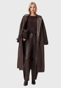 Brązowy skórzany płaszcz na ciemnobrązowym swetrze, zestawiony z dopasowanymi skórzanymi spodniami i botkami za kostkę. Strój cechuje się eleganckim, minimalistycznym designem.