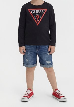 Barn iført sort langærmet GUESS-trøje med rød logo, opkrogede slidte denimshorts og røde lave sneakers med hvide snørebånd.