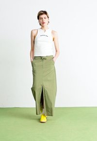 Haut sans manches blanc avec le logo "Dickies", jupe maxi olive avec fente à l'avant, et baskets jaunes sur un sol vert.