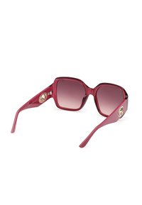 Lunettes de soleil de forme carrée avec une monture rouge translucide, des verres dégradés foncés et des logos décoratifs circulaires dorés sur les branches.